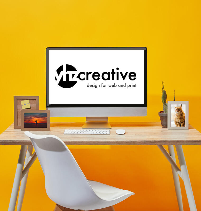 The image shows a brightly lit workspace with a vibrant yellow background. A wooden desk holds a desktop computer displaying the logo of "YHZ Creative" with the tagline "design for web and print." The desk also has a white chair, a keyboard, a mouse, two s